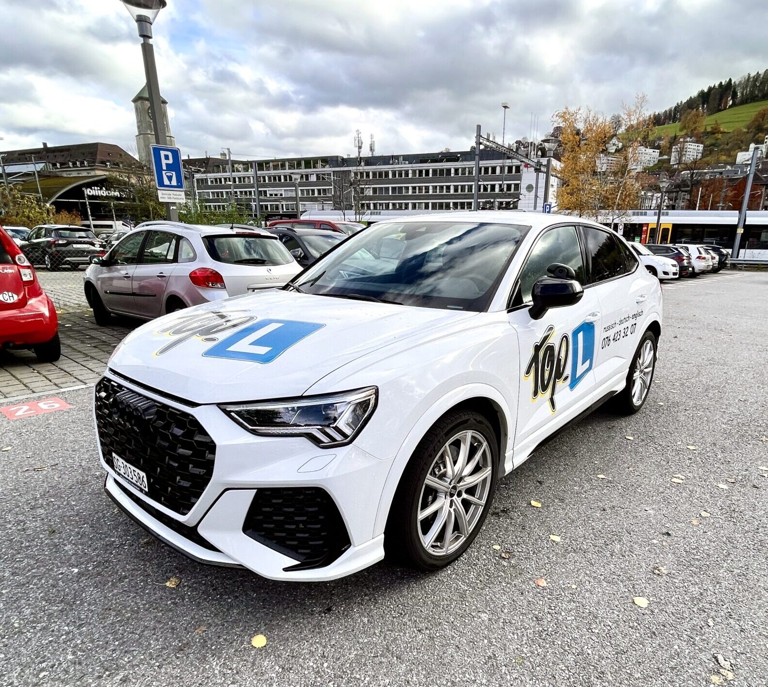 Weißes Auto mit Top L-Branding und Anfängerkennzeichen auf einem Parkplatz, umgeben von anderen Fahrzeugen. Im Hintergrund Gebäude und Bäume.
